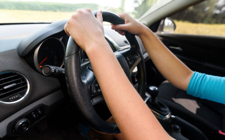 Young casual woman in blue dress driving a car, side view. Beautiful young girl at the wheel of car with black interior looking at the road